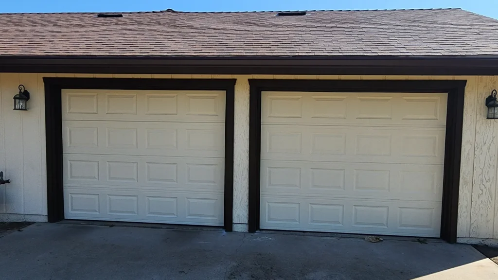 Double white garage doors on residential home with brown trim, exterior lighting, and shingled roof.