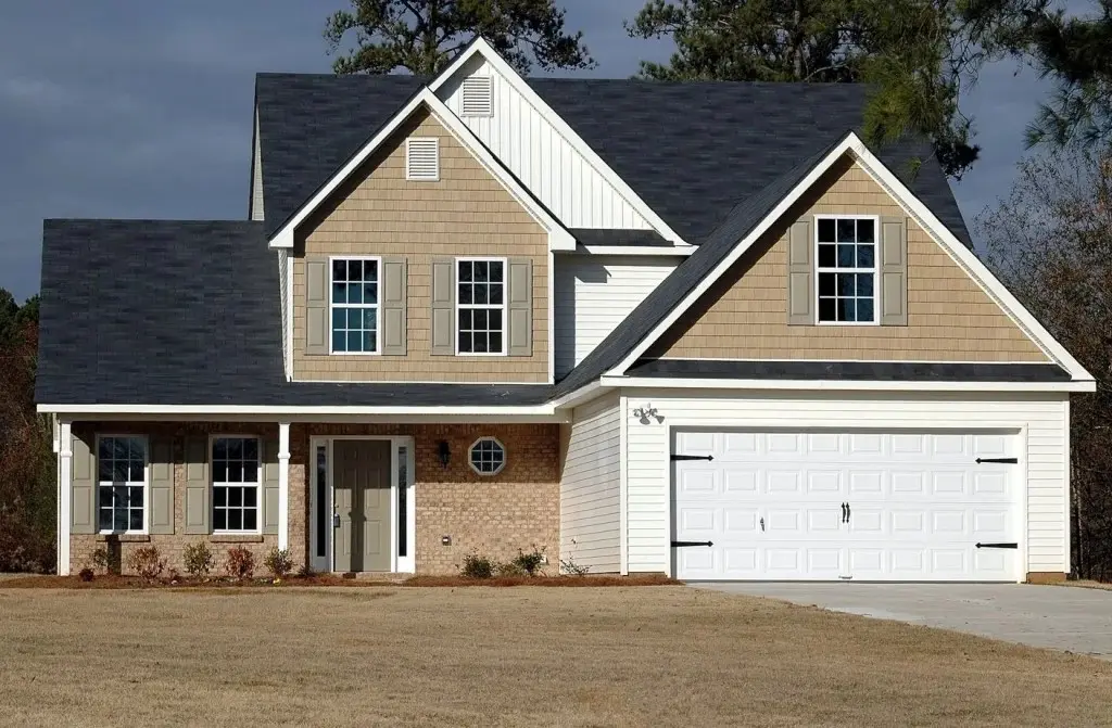 Suburban house with beige siding, dark roof, and a double garage, surrounded by a well-kept lawn and trees.