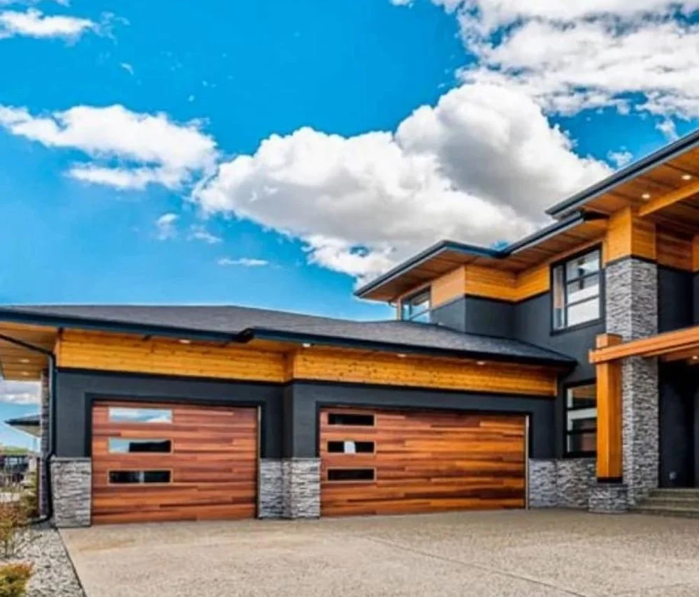 Modern two-story house with wood and stone accents, large driveway, and blue sky background.