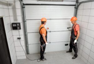 Two workers installing a garage door, wearing safety gear and holding tools, in a tiled garage.